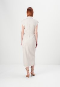 Robe beige ajustée avec des manches courtes, une silouhette à hauteur du genou et une texture lisse. Fermeture à zip au dos, portée avec des talons nu de couleur nude.