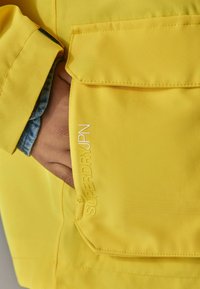 Geltona išorinė striukė su lygiu paviršiumi, turinti šoninį kišenę ir įspaustą tekstą "SUPERDRY JPN" siūlės krašte.