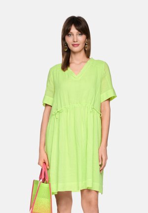 Frau trägt ein locker sitzendes limettengrünes Kleid und hält eine pink-grüne gewebte Tragetasche, steht vor einem schlichten hellen Hintergrund.