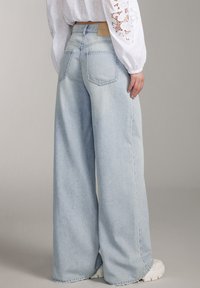 Vaqueros de pierna ancha azul claro hechos de denim, con una cintura alta, dos bolsillos traseros y un dobladillo sin terminar, combinados con una blusa blanca.