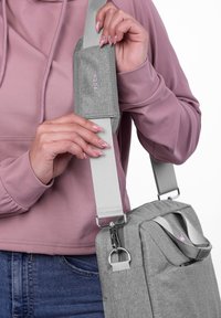 Person passt den grauen Schultergurt an der passenden grauen Stofftasche an, trägt einen rosa Kapuzenpullover und blaue Jeans, mit rosa und silbernem Nageldesign.