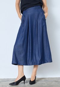 Denim A formā skaidra svārki, tumši zili, ar krokām, sānu kabatām un gludu tekstūru. Stilizēti ar melniem augstiem apaviem.