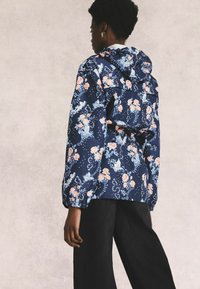 Floral-patterned, navy jacket with a hood, cinched waist, and elastic cuffs. Fabric features pink roses and blue accents, texture is smooth.