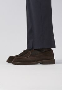 Brown suede loafers with a smooth texture, rounded toe, and contrasting stitching. Paired with navy trousers, showing a sleek, casual look.