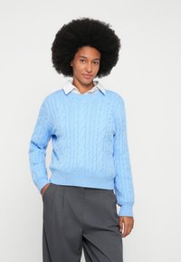 Vaaleansininen palmikkoneulepaita pyöreällä pääntiellä ja pitkillä hihoilla, yhdistetty valkoisen kaulusp paidan ja harmaiden korkeavyötäröisten housujen kanssa.