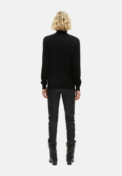 Jersey negro de cuello alto con mangas largas, combinado con pantalones de cuero negros. Modelo con botas de vaquero negras, visto desde atrás.