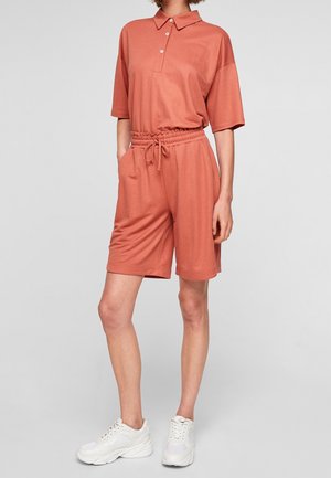 Personne portant un polo corail ample et un short à cordon assorti, associé à des baskets blanches épaisses, debout devant un fond uni.