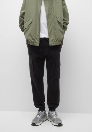 Veste bomber vert olive avec des poignets élastiques, chemise blanche en dessous, pantalon cargo noir avec poches latérales, et baskets grises.