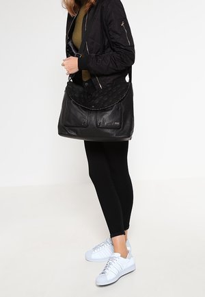 Femme portant une veste noire, des leggings noirs, des baskets blanches, portant un grand sac à main en cuir noir avec rabat et poches avant.