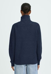 Marine gebreide trui met hoge kraag, gestructureerd patroon en ribbelzoom. Oversized pasvorm met lange mouwen. Draagt bij lichte blauwe jeans.