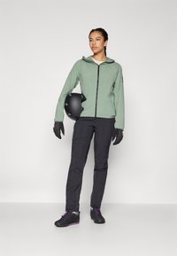 Helle grüne Kapuzenjacke mit Reißverschluss, schwarze Hose, schwarzer Helm und Handschuhe. Auffällige Details sind lila Akzente an den Schuhen.