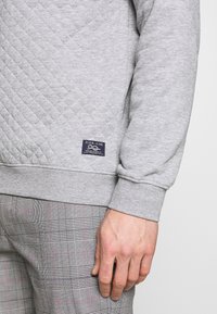 Sweat-shirt matelassé gris avec poignets et ourlet côtelés, avec une petite étiquette marine indiquant "PIER ONE" près de la taille. Associé à un pantalon à carreaux.