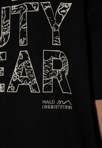 Svart tyg med stora vita konturerade bokstäver som visar abstrakta kartmönster och mindre text "HALO 196819721981" nedanför.