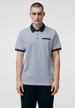 Młody mężczyzna w białej koszulce polo w wzory z granatowym kołnierzem i mankietami, stojący na tle jasnego, jednolitego tła.