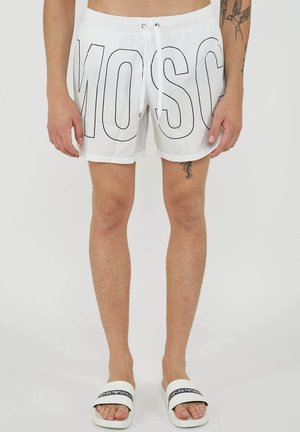 Homme portant un short blanc avec de grandes lettres noires en contour et des sandales blanches avec le logo noir Emporio Armani.