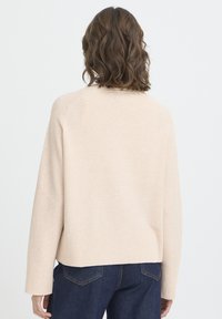 Pulover bej deschis cu textură reiată, mâneci largi și croială lejeră. Asortat cu pantaloni din denim închis, văzut din spate.