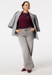 Grijze geruite blazer, bordeauxkleurig graphic t-shirt, lichtgrijze flared jeans en bordeauxkleurige flats. Model poseert met de handen in de zakken. Eenvoudige achtergrond.