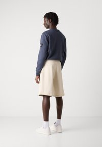 Navy sweatshirt with a logo patch and beige sweat shorts. The outfit includes white sneakers and visible ankle socks. Smooth cotton texture.