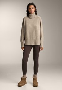 Pulover bej cu guler înalt, asociat cu colanți maro închis și cizme scurte cu blăniță bej, pe un fundal neutru.