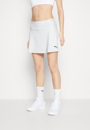 Kvinde iført hvid atletisk skort, hvid sports-bh, hvide Puma-strømper og hvide sneakers, stående mod en ensartet lys baggrund.