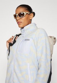 Giacca in fleece azzurro chiaro con motivi gialli, collo alto e tasca sul petto. Cinghia dello zaino nera visibile sulla spalla. Occhiali da sole marroni.