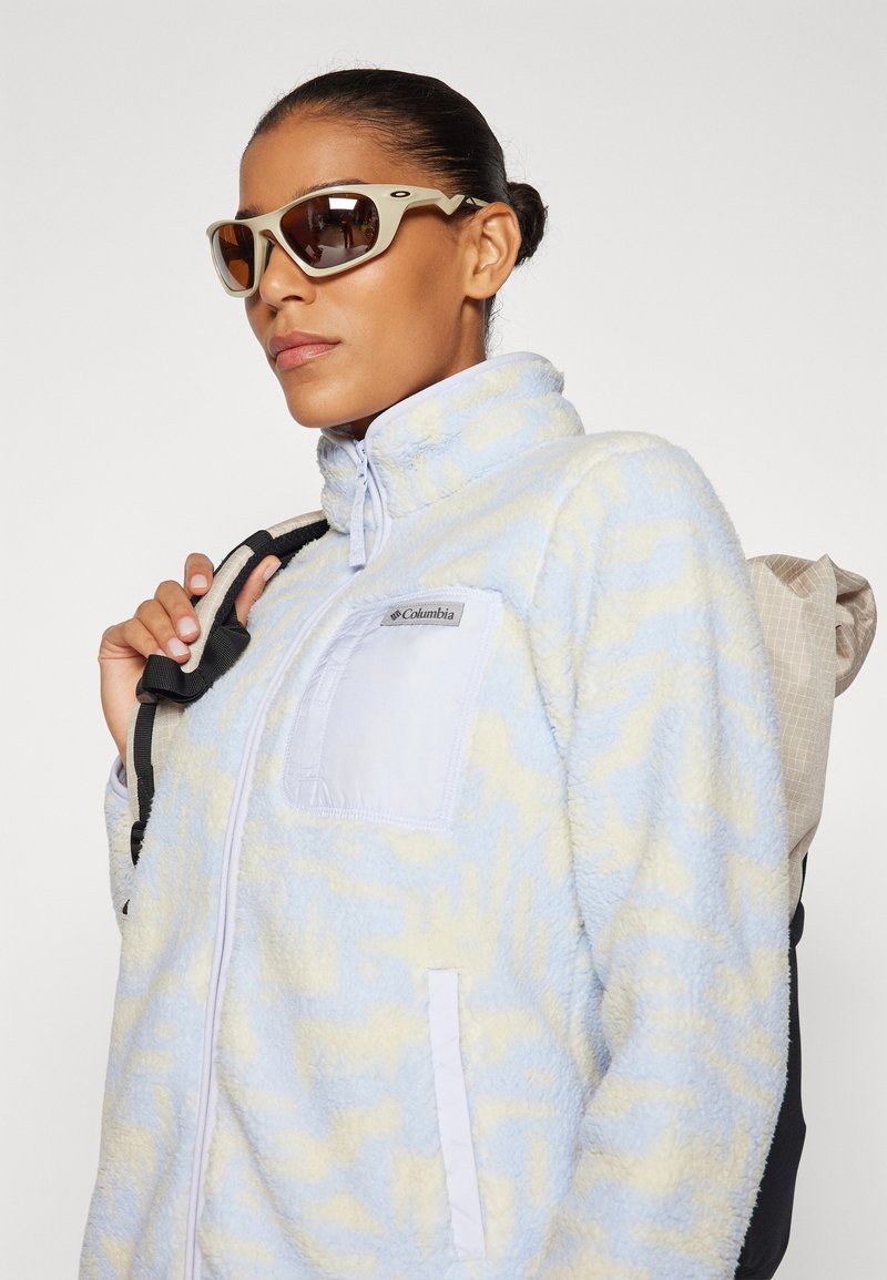 Lichtblauwe fleece-jack met gele patronen, hoge kraag en een borstzak. Zwarte rugzakband zichtbaar over de schouder. Bruine zonnebril.