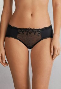 Black lingerie brief featuring a sheer lace front with polka dots, scalloped edges, and decorative bows on the waistband. Smooth fabric back.