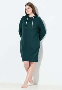 Mujer de pie descalza contra una pared blanca, vestida con un vestido oscuro verde con capucha y cordones blancos, con las manos detrás de la espalda.