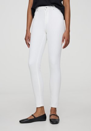 Jeans bianchi slim-fit con vita alta, realizzati in tessuto denim. Dotati di chiusura frontale con bottone e cuciture laterali discrete. Indossati con ballerine nere.