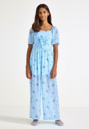 Pige stående iført en lyseblå blomsterkjole med pufærmer og bindebånd foran, kombineret med lysefarvede mesh slip-on sko.