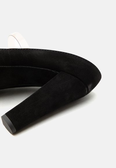 Μαύρο σουέντ γυναικείο παπούτσι με ψηλό τακούνι και χοντρό τακούνι, με ελαφρώς στρογγυλεμένη μύτη, εμφανιζόμενο σε απλό λευκό φόντο.