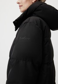 Naine kannab musta puffer-jopet kõrge krae ja kapuutsiga, külgvaade, kus on nähtav "LeGer" logo varrukal.