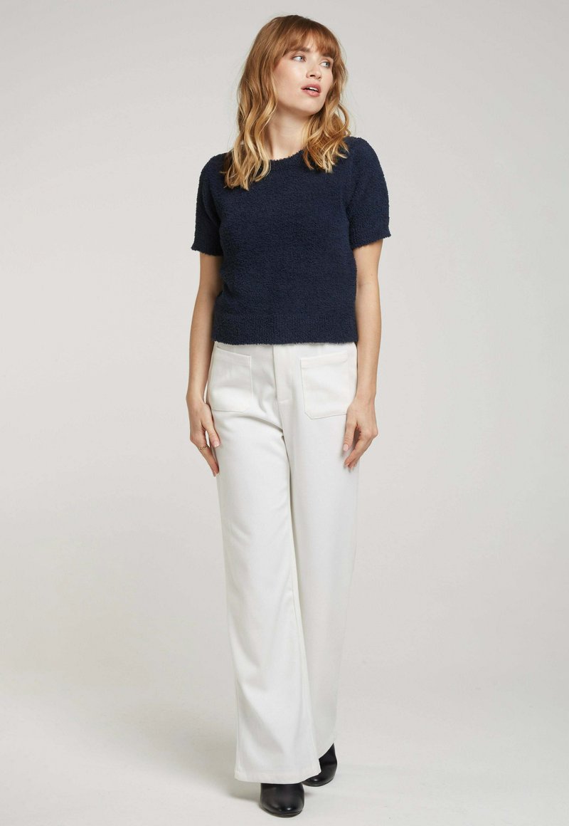 Pull en maille texturée bleu marine à manches courtes associé à un pantalon blanc à jambes larges et des bottines noires. Design simple sans motifs visibles.