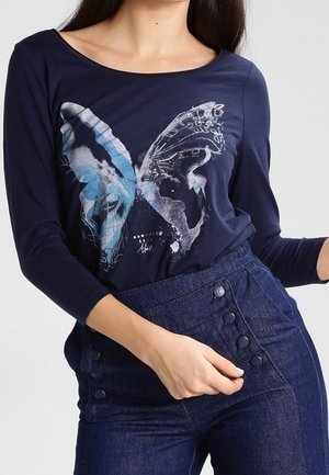 Femme portant une chemise bleu marine à manches longues avec un grand papillon bleu et blanc graphique, et un pantalon bleu foncé taille haute à boutons.