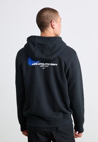Mand iført sort hættejakke med blå Nike-logo og hvid tekst "Nike Athletic Dept Just Do It", vender væk fra en ensfarvet baggrund.