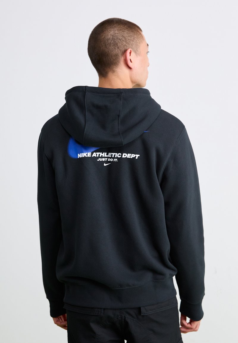 Mand iført sort hættejakke med blå Nike-logo og hvid tekst "Nike Athletic Dept Just Do It", vender væk fra en ensfarvet baggrund.