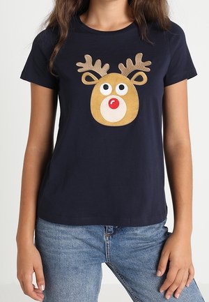 Vrouw die een donkerblauw T-shirt draagt met een cartoonrendiergezicht met grote ogen en een rode neus, gecombineerd met blauwe spijkerbroek.