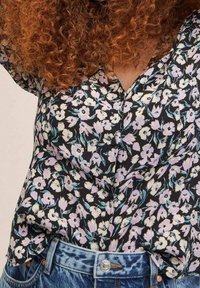 Blumenbluse mit schwarzem Hintergrund, die mit weißen und lila Blumen verziert ist, im V-Ausschnitt-Design und aus weichem Stoff, kombiniert mit hochtaillierten Jeans.