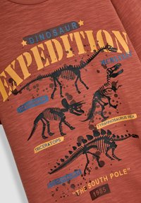 Rode graphic t-shirt met dinosaurusskeletten: Tyrannosaurus Rex, Triceratops, Stegosaurus, met tekst en kleurrijke accenten.