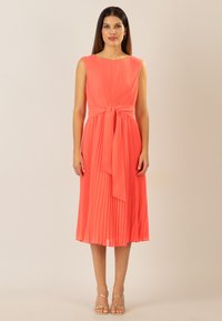 Korallenfarbenes plissiertes Kleid mit tailliertem Oberteil, rundem Ausschnitt und Taillengürtel. Glatter Stoff, knielanger Saum und minimales Zubehör.