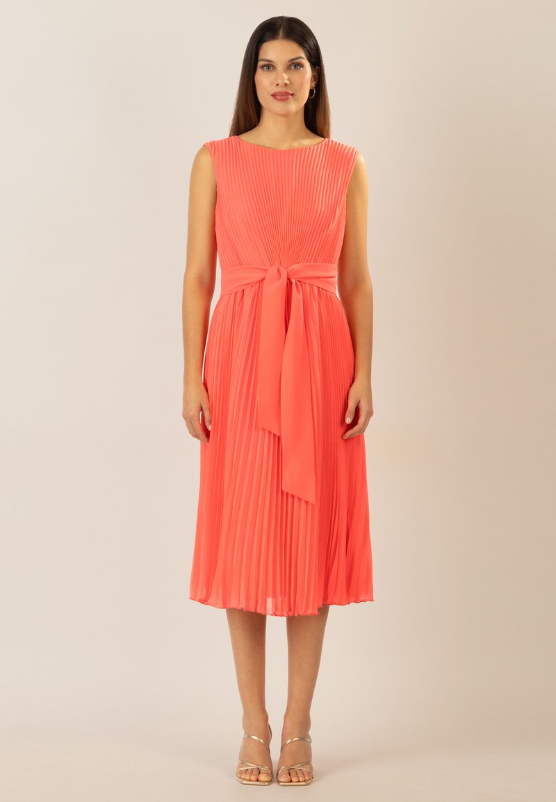 Korallenfarbenes plissiertes Kleid mit tailliertem Oberteil, rundem Ausschnitt und Taillengürtel. Glatter Stoff, knielanger Saum und minimales Zubehör.
