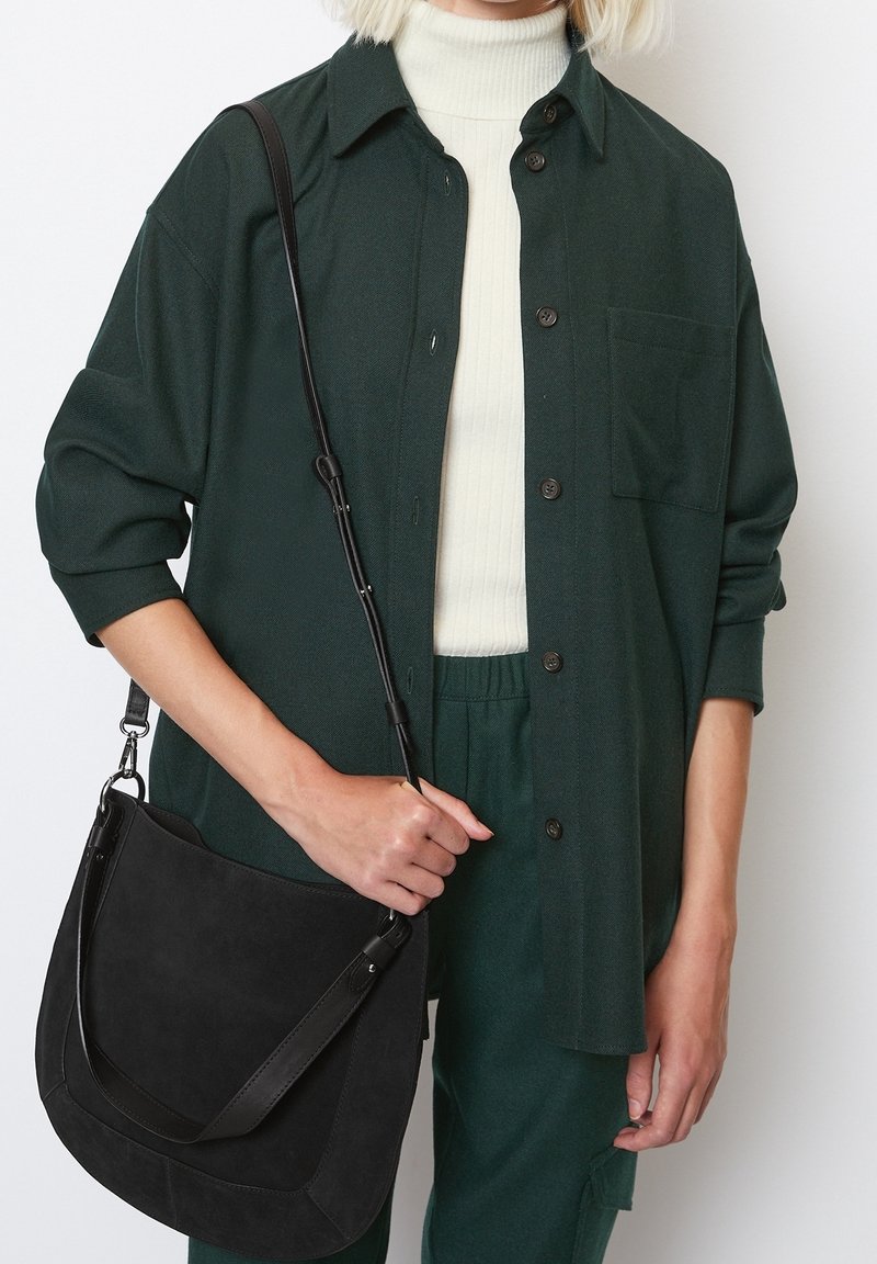 Personne portant une chemise boutonnée vert foncé et un pantalon, un col roulé côtelé blanc, tenant un sac à bandoulière en daim noir avec une sangle réglable.