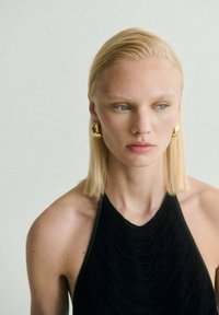 Černý top s obdélníkovým výstřihem vyrobený z texturované tkaniny, s hladkým výstřihem. Modelka má rovné, blond vlasy a zlaté, kulaté náušnice.