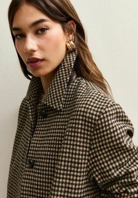 Abrigo con patrón de pata de gallo en beige y negro, con un cuello alto, grandes botones y detalles de cuero en los hombros. Tejido suave y texturizado.