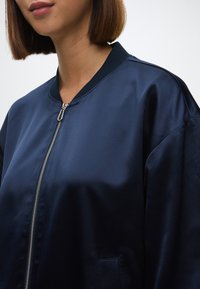 Jūras spēku iznīcinātāja jaka ar gludu satīna tekstūru, ribotu apkakli un sudraba rāvējslēdzēju. Iekļauj sānu kabatu un brīvu piegriezumu.