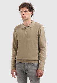 Beige Langarm-Polohemd mit strukturiertem Strickdesign, Drei-Knopf-Leiste sowie geripptem Kragen und Bündchen, kombiniert mit grauen Jeans.