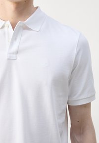 Homme portant un polo blanc uni à manches courtes avec deux boutons visibles et un logo brodé subtil sur la poitrine.