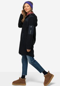 Cappotto in pile nero con cappuccio, applicazione trapuntata sulle spalle; abbinato a jeans blu e stivaletti marroni alla caviglia. Il modello indossa un berretto a righe viola.