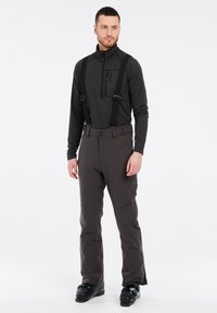 Herrenmode mit einem schwarzen Langarmoberteil mit einer Reißverschlusstasche und grauen Schneehosen mit Hosenträgern, Stiefeln und minimalem Branding. Glattes, strapazierfähiges Material.