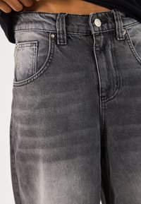 Vaqueros de mezclilla gris con un acabado desgastado. Cuenta con cinco bolsillos, cierre de botones y herrajes metálicos. La tela tiene una textura lisa y resistente.